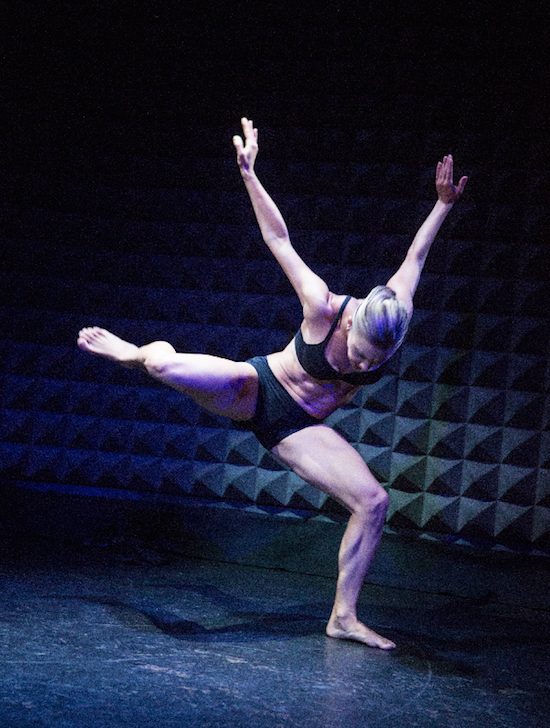 A female dancer in a bra top and shorts balances on one leg with her standing leg bent. Her torso and head face the floor as her arms extend behind her.